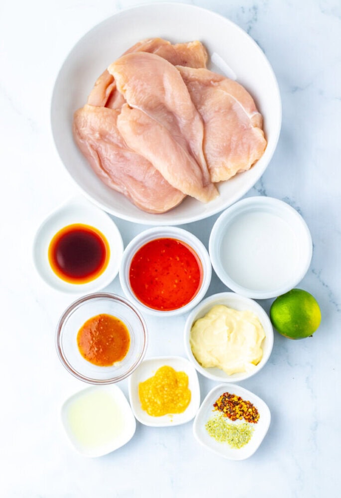 Raw chicken breasts in a bowl surrounded by small bowls of various sauces, seasonings, lime, and liquid ingredients on a white surface.