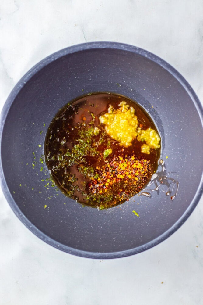 A black bowl containing a mixture of oil, minced garlic, dried herbs, red pepper flakes, and possibly vinegar on a light surface.