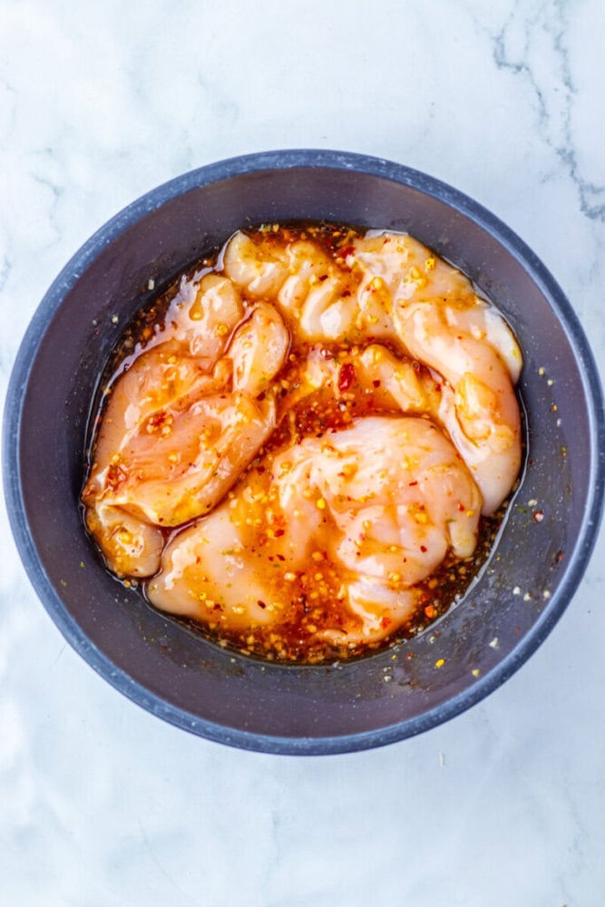 Raw chicken breasts marinating in a bowl with a mixture of oil and spices on a white marble surface.