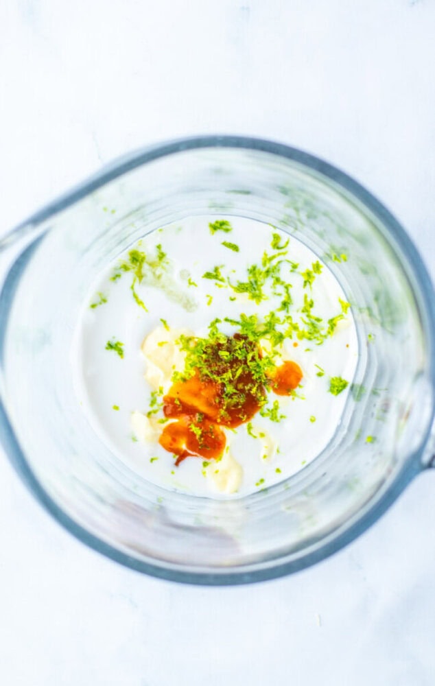 A glass bowl containing a mixture of white liquid, lime zest, a brown sauce, and a dollop of white paste on a white surface.