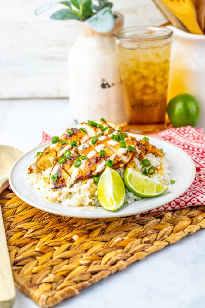 A plate of grilled chicken over rice, topped with sauce and chopped green onions, served with lime wedges. A glass of iced drink and a potted plant are in the background.