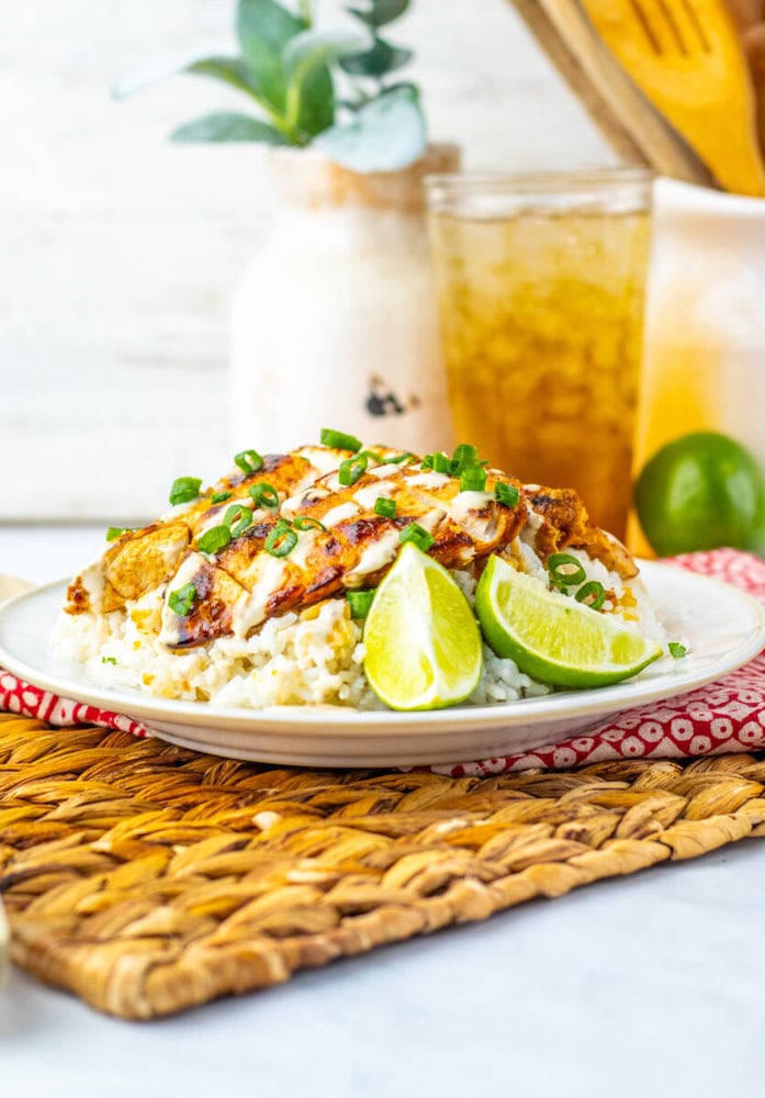 Grilled chicken breast slices on rice, garnished with chopped green onions and lime wedges, served on a plate with a glass of iced tea in the background.