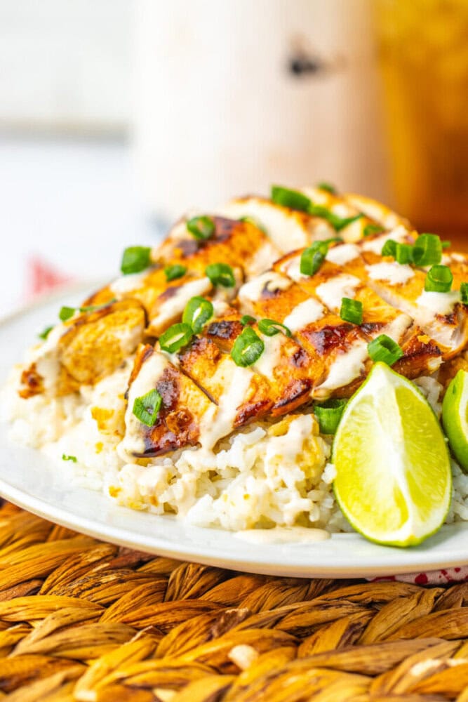 Grilled chicken breast slices with creamy sauce and chopped green onions served over rice, garnished with fresh lime wedges on a white plate.