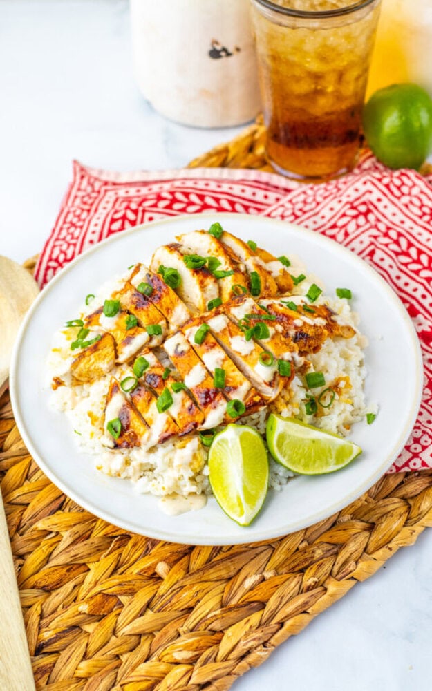 A plate of sliced grilled chicken on a bed of rice, garnished with chopped green onions and lime wedges, sits on a woven mat with a red patterned napkin nearby.