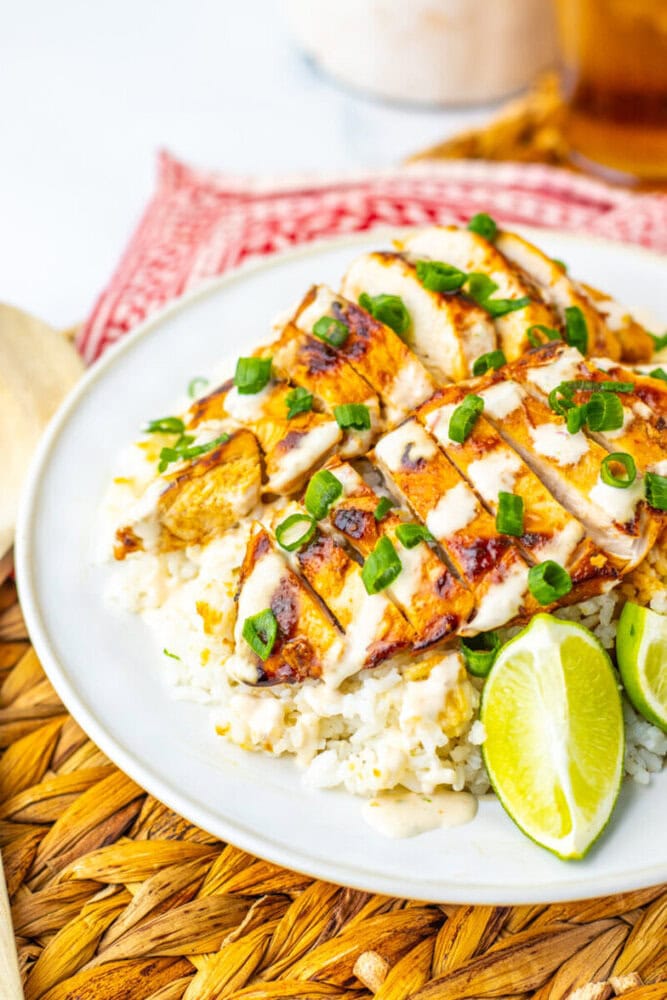 Sliced grilled chicken with sauce and chopped green onions served over rice, garnished with lime wedges on a white plate.