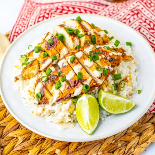 A plate of grilled chicken slices on a bed of rice, topped with chopped green onions and sauce, with lime wedges on the side. A wooden spoon and red patterned napkin are nearby.