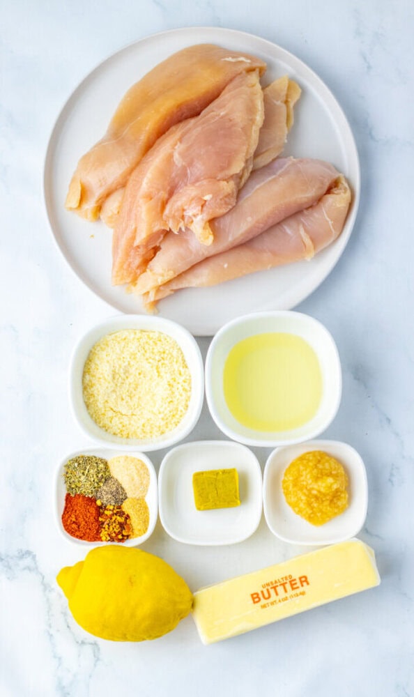 Raw chicken breasts on a plate, surrounded by small bowls of breadcrumbs, oil, spices, bouillon, minced garlic, a lemon, and a stick of unsalted butter on a white background.