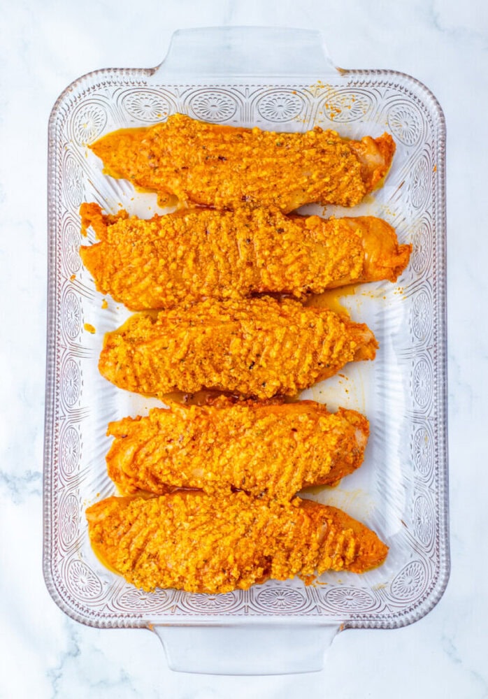 Four breaded fish fillets arranged in a single layer on a clear glass baking dish, ready to be cooked.