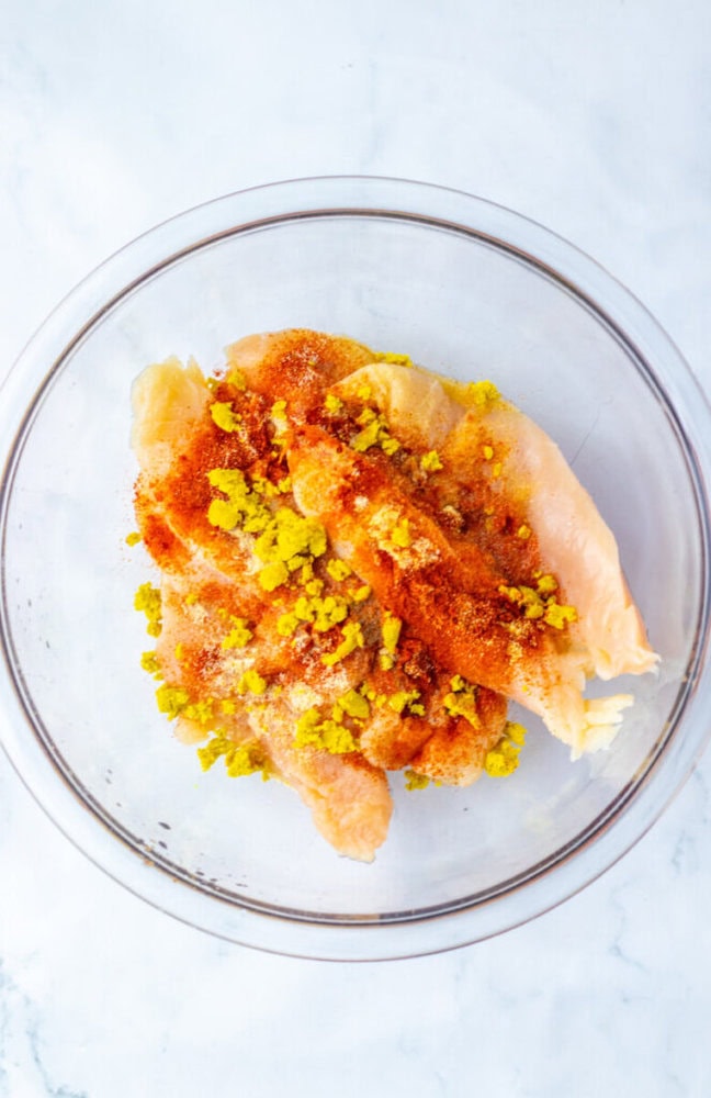 Raw chicken breast fillets in a glass bowl, topped with spices and what appears to be chopped yellow pickles, on a white surface.