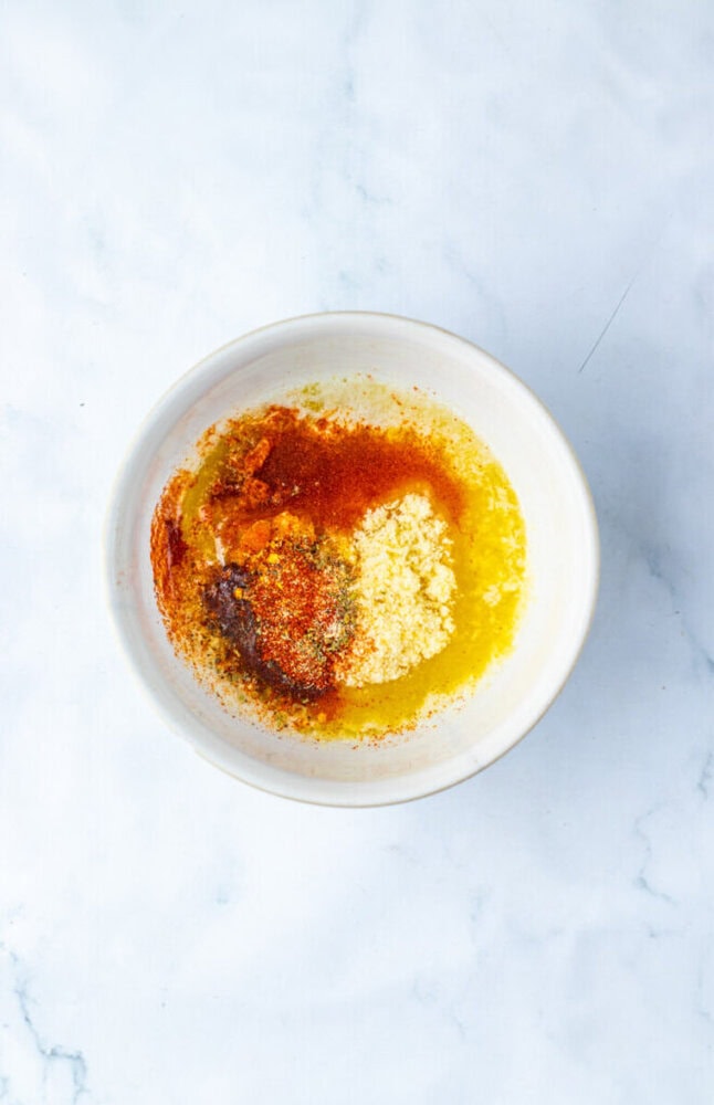 A white bowl containing melted butter, grated parmesan, dried herbs, and paprika on a light marble surface.