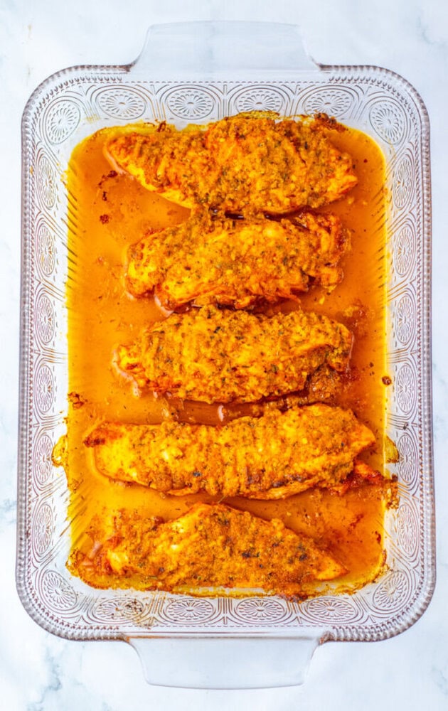 Five baked chicken breasts coated with spices and sauce are arranged in a rectangular glass baking dish on a white surface.