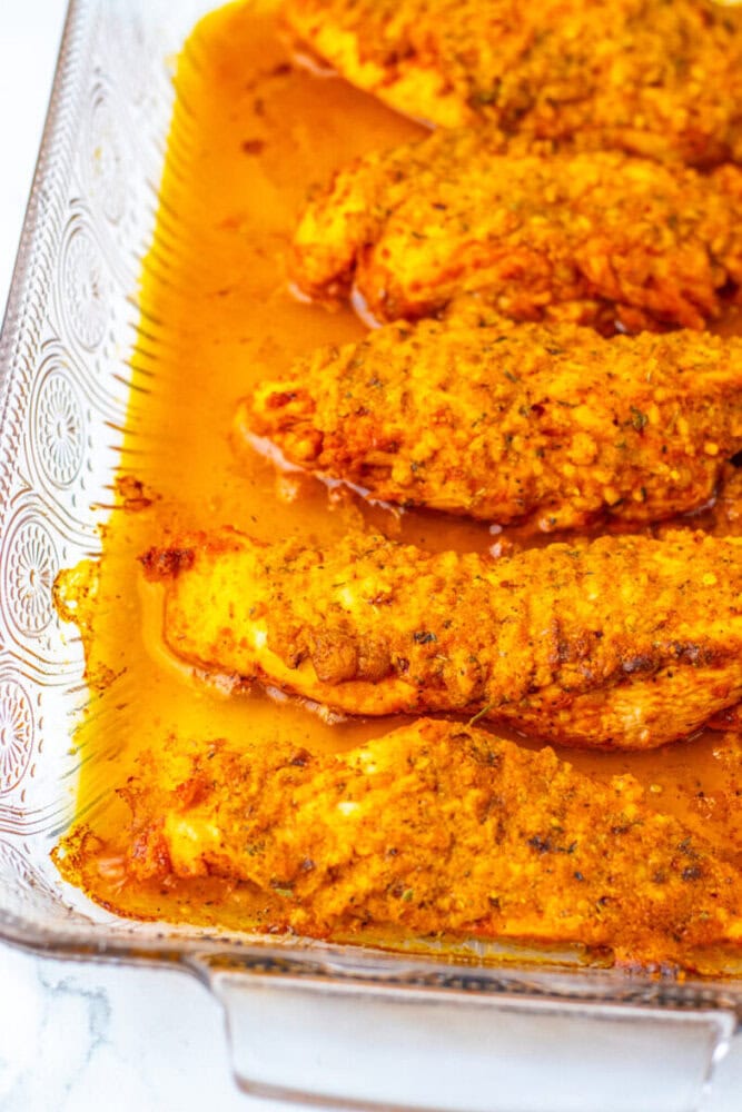 Five seasoned and baked chicken breasts in a glass dish, covered with a yellow-orange sauce.