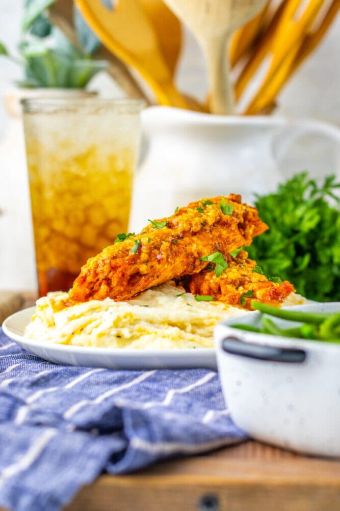 Two pieces of baked or grilled fish with tomato-based sauce served over mashed potatoes, with a drink and green vegetables in the background.