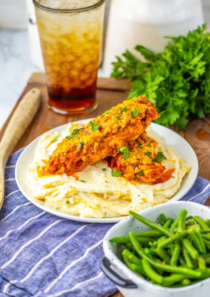 Plate with mashed potatoes and two pieces of breaded chicken, garnished with herbs; green beans in a small colander and a glass of iced tea in the background.