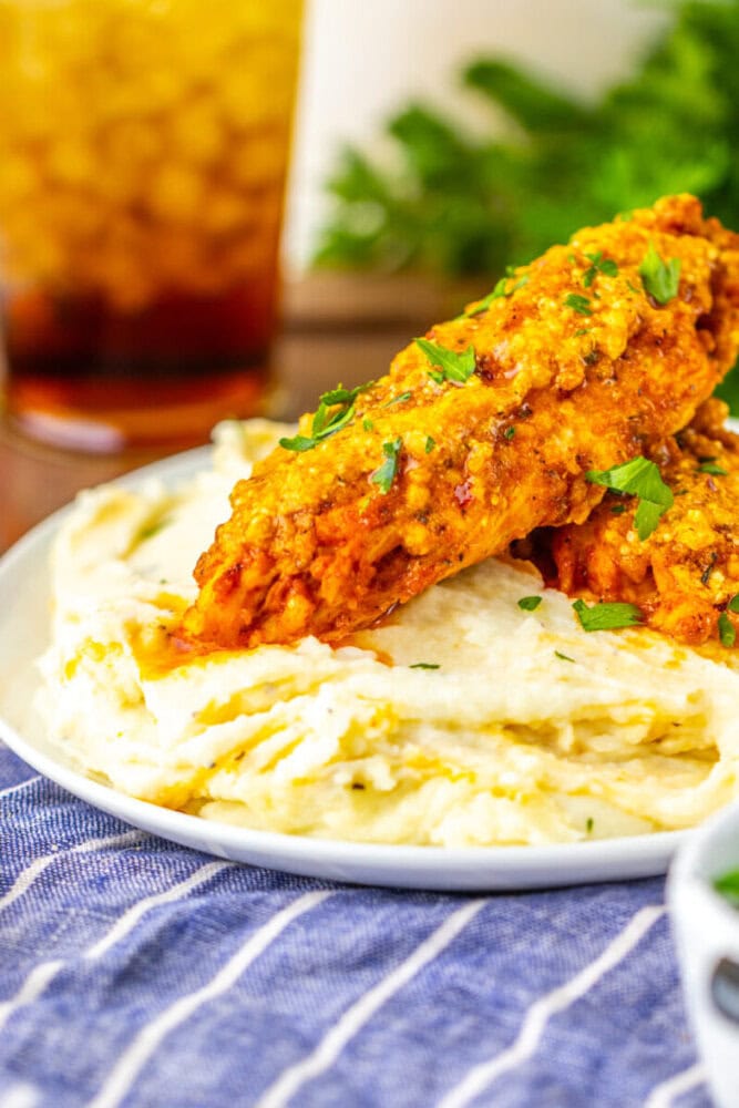 Two breaded chicken tenders coated in sauce served on a bed of mashed potatoes, garnished with chopped herbs.