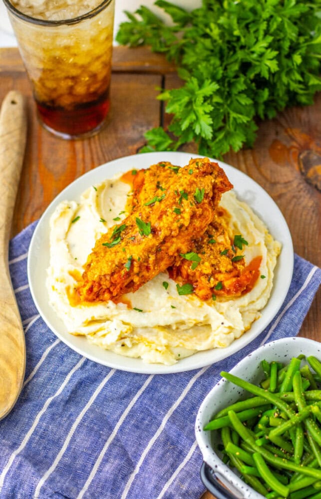 Plate of mashed potatoes topped with two pieces of fried chicken, garnished with parsley, next to a bowl of green beans and a glass of iced drink on a wooden table.