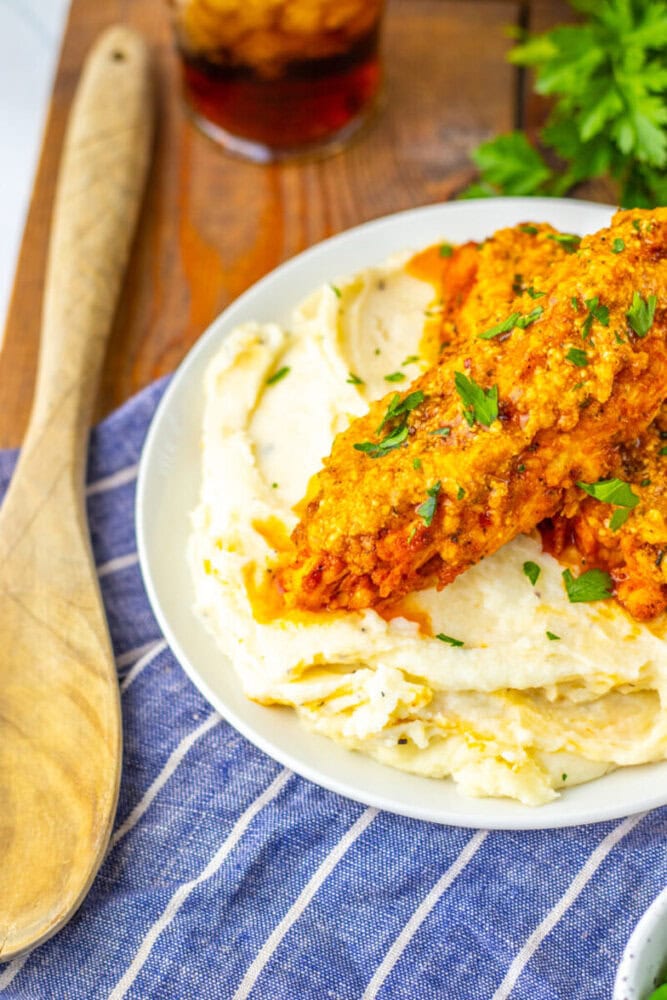 A plate of mashed potatoes topped with two pieces of breaded chicken in orange sauce, garnished with chopped parsley.