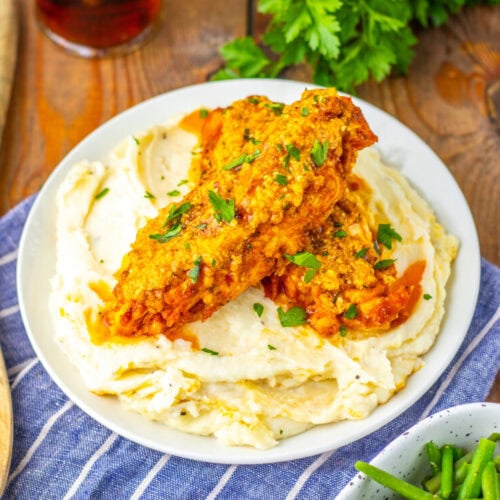 Two pieces of fried chicken served on a bed of mashed potatoes, garnished with chopped herbs, on a white plate with a wooden spoon and green beans nearby.