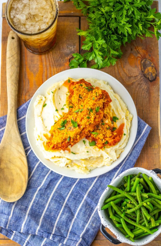 A plate of mashed potatoes topped with two pieces of fried chicken, garnished with parsley, next to green beans, a drink with ice, and a bunch of fresh parsley on a wooden table.