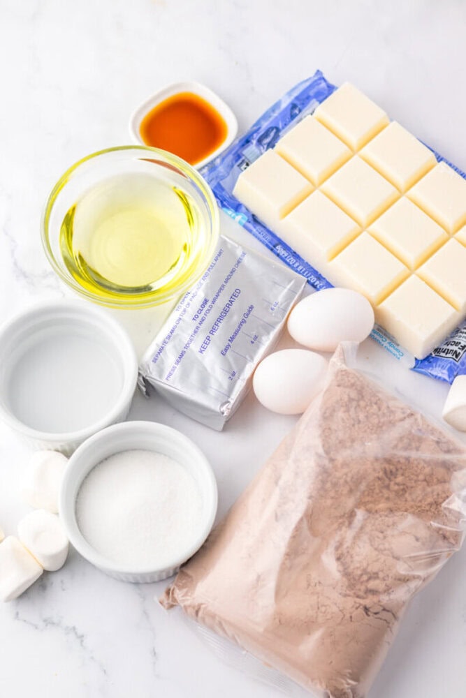 Ingredients for baking laid out on a white surface, including white chocolate, eggs, oil, sugar, cocoa powder mix, marshmallows, vanilla extract, and packaged cream cheese.