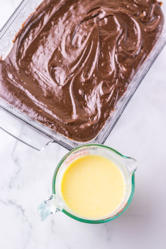 A glass baking dish filled with chocolate batter sits beside a measuring cup containing yellow batter on a marble countertop.