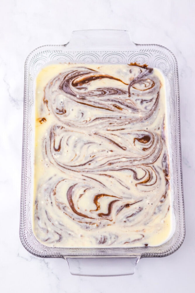 A glass baking dish filled with marbled cheesecake batter, swirled with chocolate, on a white marble surface.