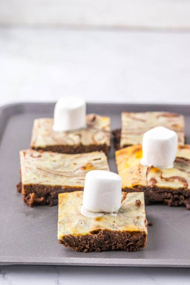 Four chocolate and cream cheese swirl brownie squares on a baking sheet, each topped with a single large marshmallow.