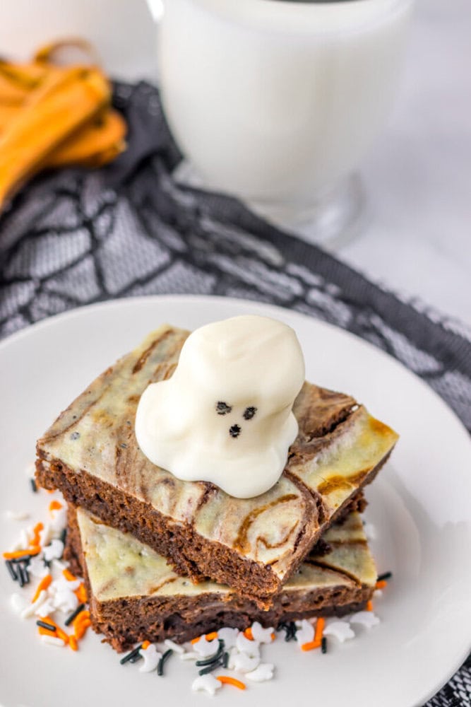 Two brownies with a marbled top are stacked on a white plate, topped with a ghost-shaped white candy and surrounded by Halloween-themed sprinkles. A glass of milk is in the background.