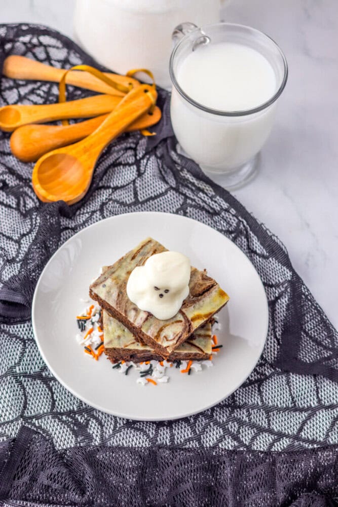 A plate with two marbled brownies topped with whipped cream shaped like a ghost, next to a glass of milk and measuring spoons on a black lace cloth.