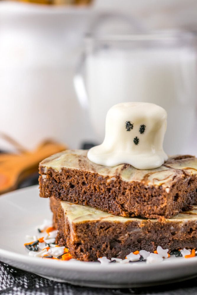 Two marble brownies stacked on a plate, topped with a white chocolate ghost-shaped decoration with black eyes and a mouth. A glass of milk is in the background.