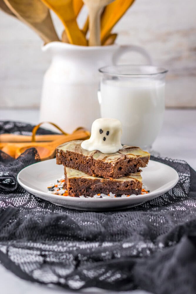 Two brownies topped with a white chocolate ghost decoration on a white plate, with a glass of milk and a pitcher in the background.