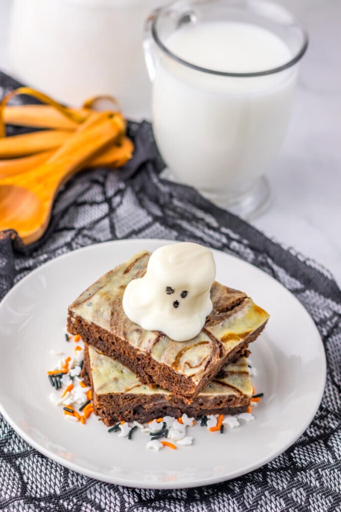 A plate with two marbled brownies topped with a ghost-shaped dollop of frosting, next to a glass of milk and measuring spoons, on a black lace cloth.