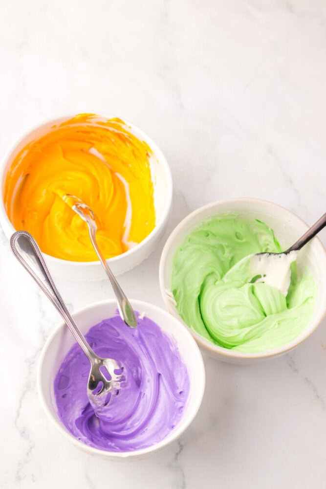 Three bowls of colored frosting—yellow, green, and purple—each with a spoon, sit on a white marble surface.