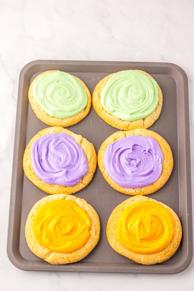 A baking tray with six round cookies, each topped with swirled frosting in light green, purple, or yellow, arranged in two rows on a marble surface.