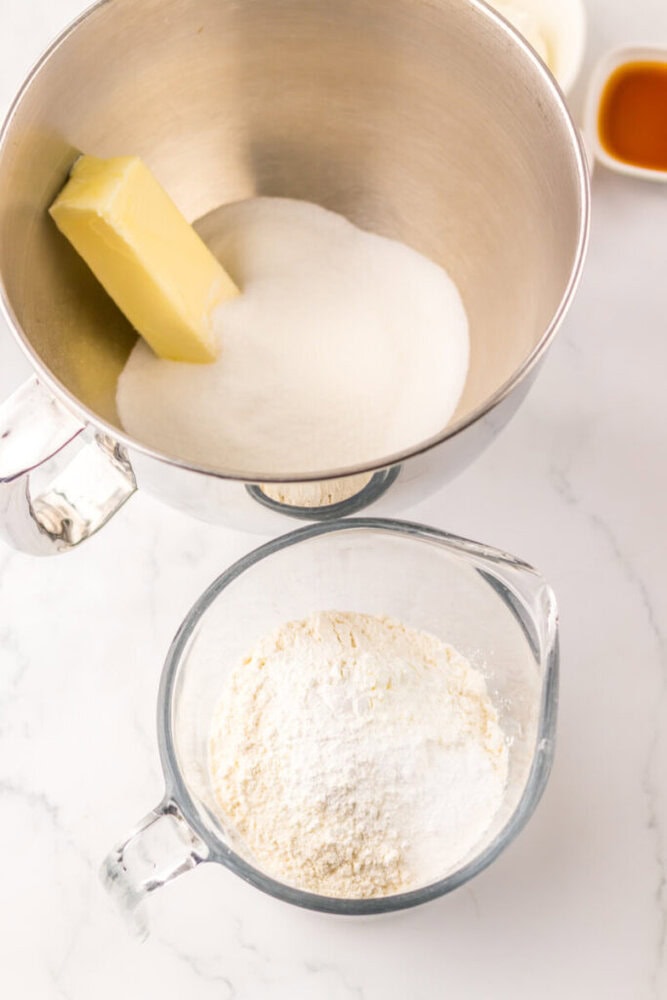 A metal mixing bowl with butter and sugar next to a glass measuring cup containing flour and baking powder on a white surface.