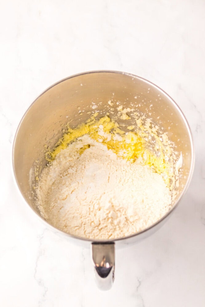 A metal mixing bowl containing creamed butter and sugar with flour added on top, ready to be mixed on a white background.