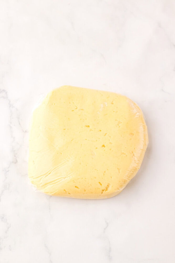 A block of yellow dough wrapped in plastic wrap rests on a white marble surface.