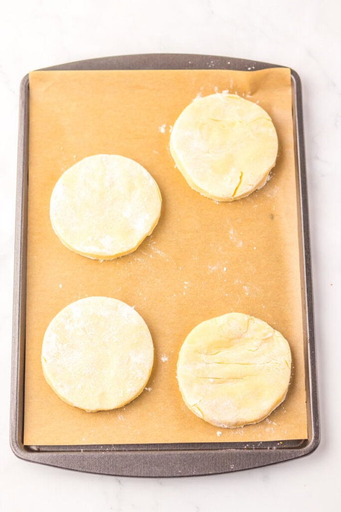 A baking sheet lined with parchment paper holds four uncooked, round biscuit dough pieces lightly dusted with flour.