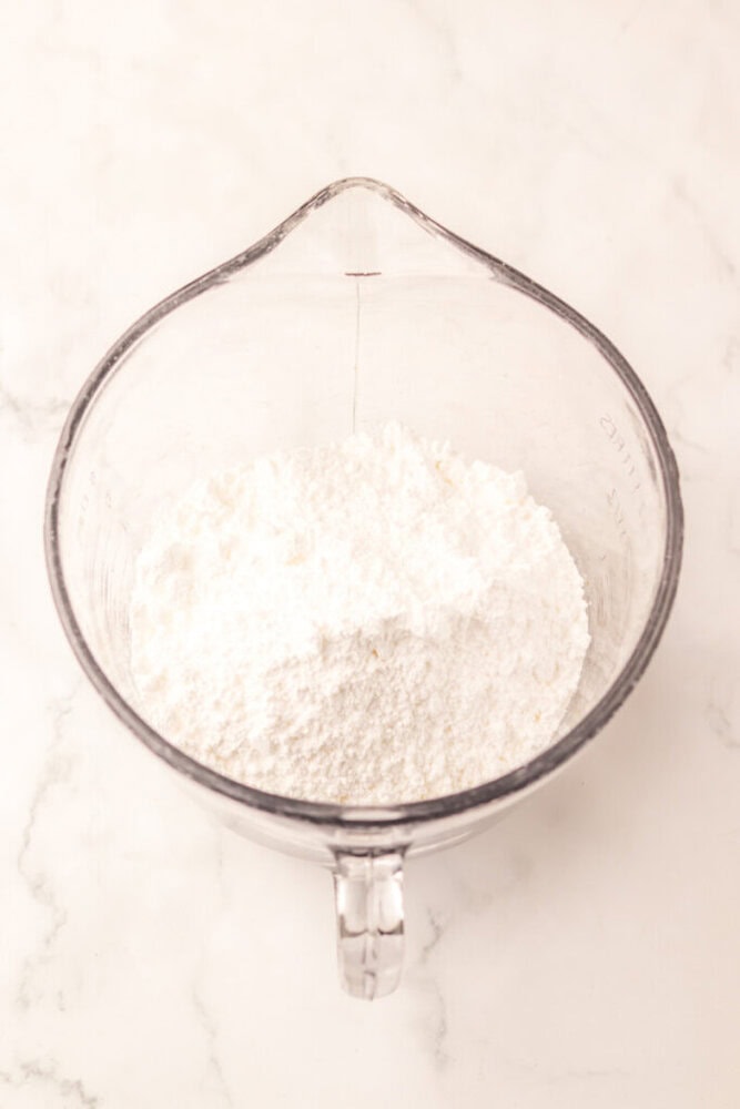 A glass mixing bowl filled with a mound of white powdered sugar on a white marble surface.