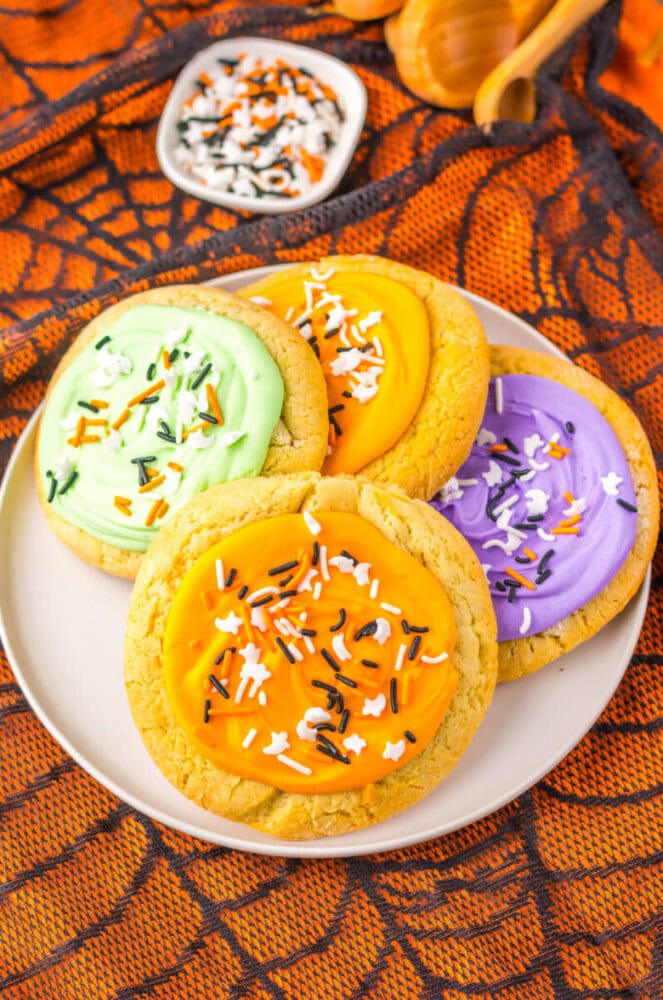 A plate with four frosted sugar cookies in green, orange, and purple colors, topped with Halloween-themed sprinkles on an orange spiderweb-patterned tablecloth.