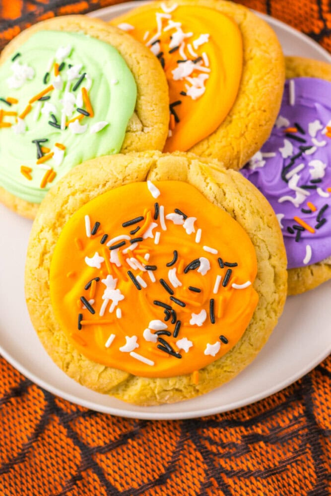 A plate of sugar cookies with orange, green, and purple frosting, decorated with black, white, and orange sprinkles, on a Halloween-themed cloth.