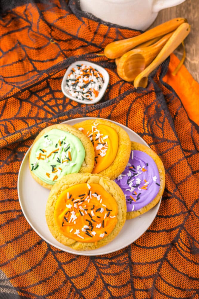 A plate of frosted sugar cookies with green, orange, and purple icing, topped with Halloween sprinkles, on an orange spiderweb-patterned cloth.