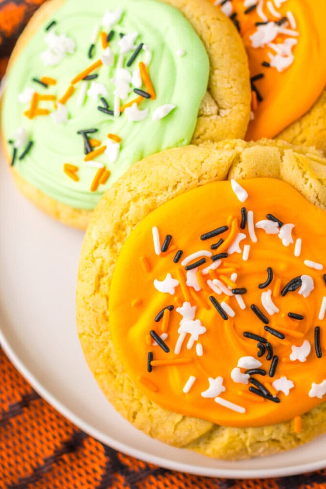 Three large cookies on a plate, topped with orange and green frosting, and decorated with black, white, and orange sprinkles.