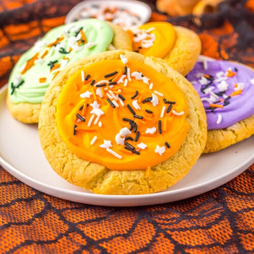 A plate with frosted sugar cookies decorated with orange, green, and purple icing and sprinkles, set on an orange spiderweb-patterned cloth.