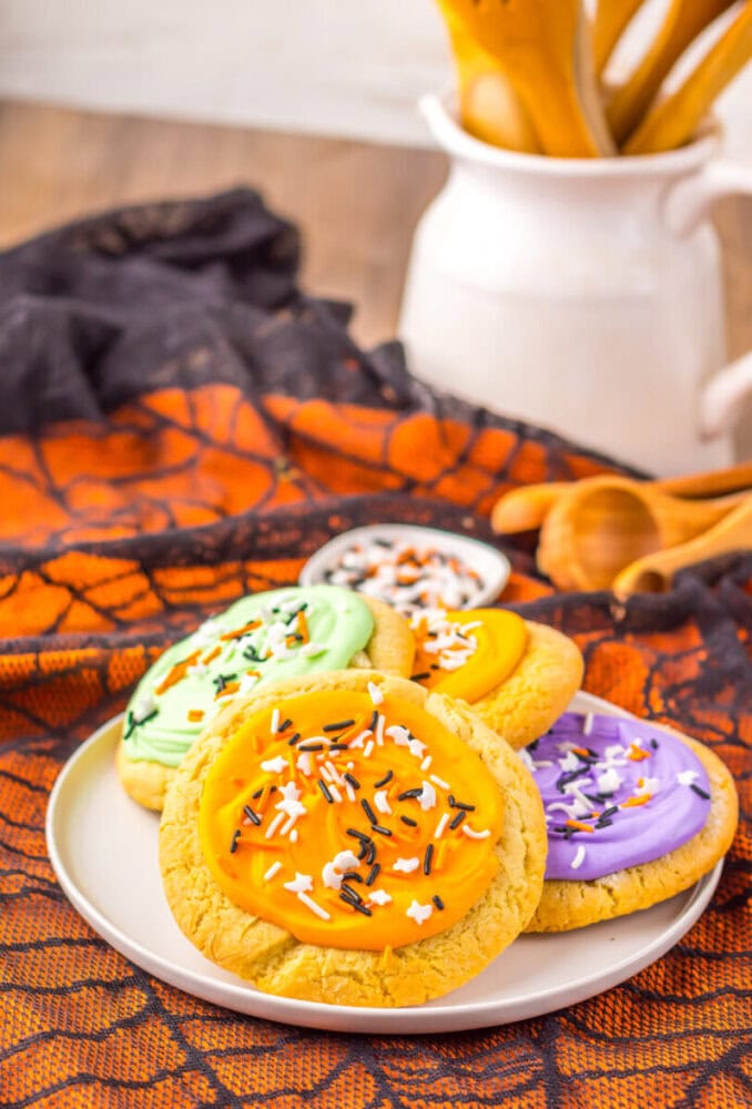 A plate of frosted sugar cookies with orange, green, yellow, and purple icing and Halloween-themed sprinkles, set on an orange and black cloth with baking utensils in the background.