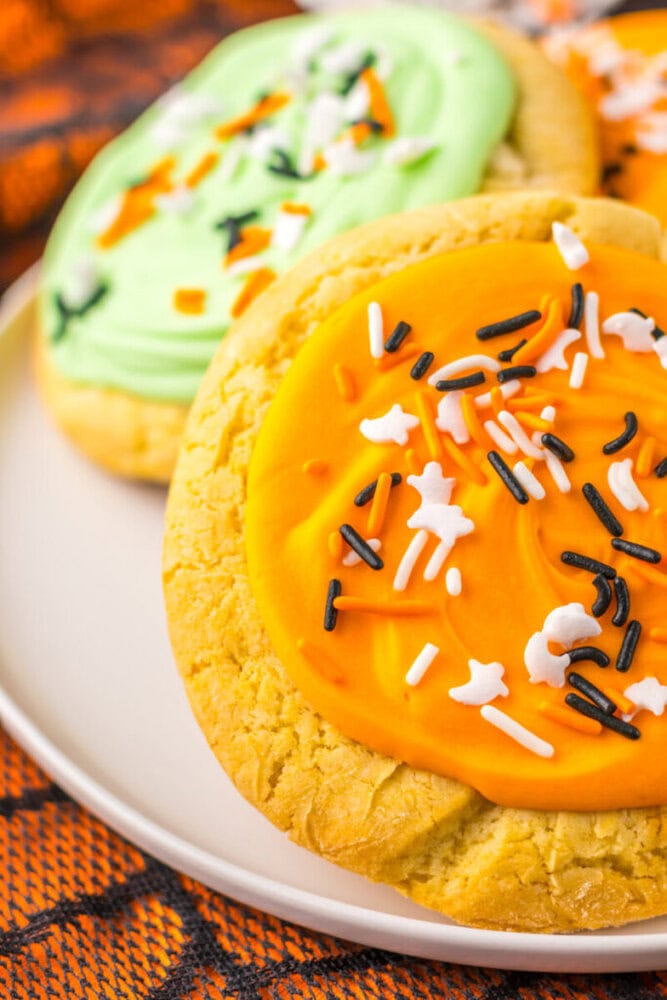 Close-up of frosted sugar cookies with orange and green icing, topped with black, white, and orange sprinkles, on a white plate.