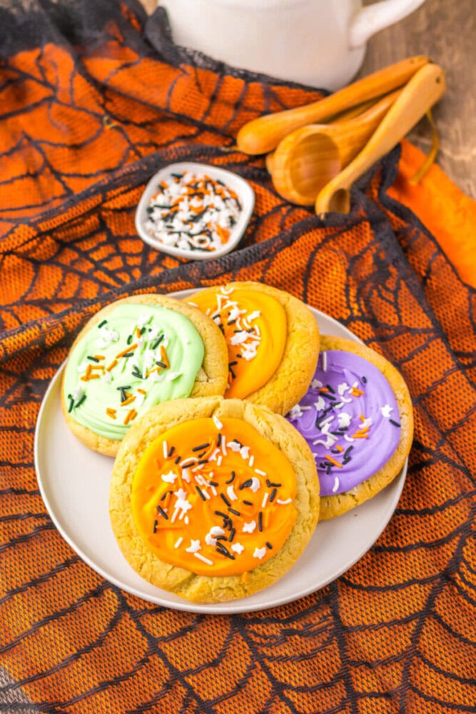 A plate of cookies with green, orange, and purple frosting topped with Halloween-themed sprinkles, set on an orange spiderweb-patterned cloth.