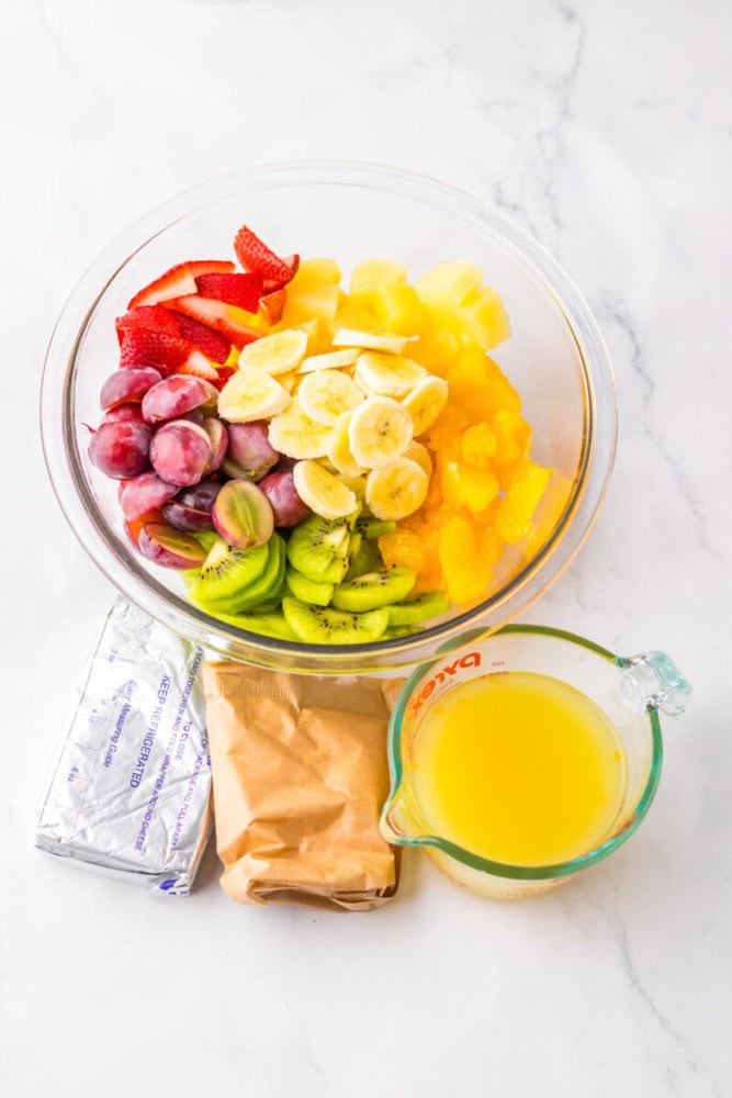 A glass bowl filled with sliced strawberries, grapes, kiwi, bananas, pineapple, and oranges sits next to cream cheese, a brown packet, and a measuring cup of orange juice.