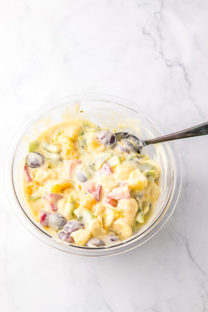 A glass bowl filled with mixed fruit salad coated in creamy dressing, with a spoon resting inside, placed on a white marble surface.