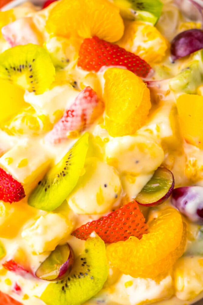 A close-up of a fruit salad with strawberries, kiwi, mandarin oranges, grapes, pineapple, and creamy dressing.